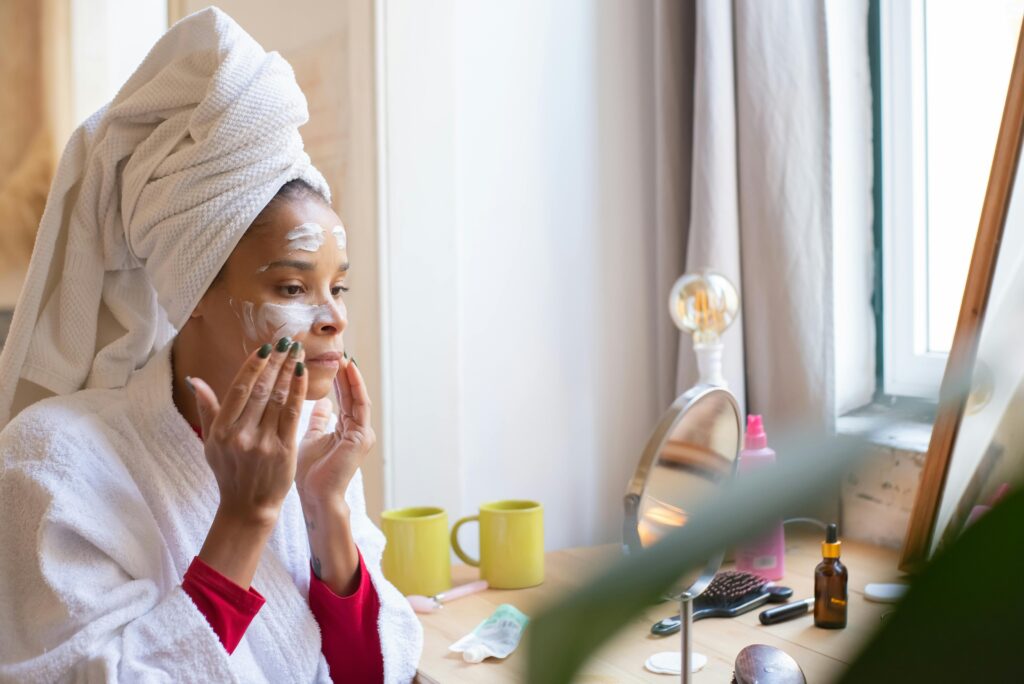 A woman applies face cream in a relaxing home setting, dressed in a bathrobe and head towel.